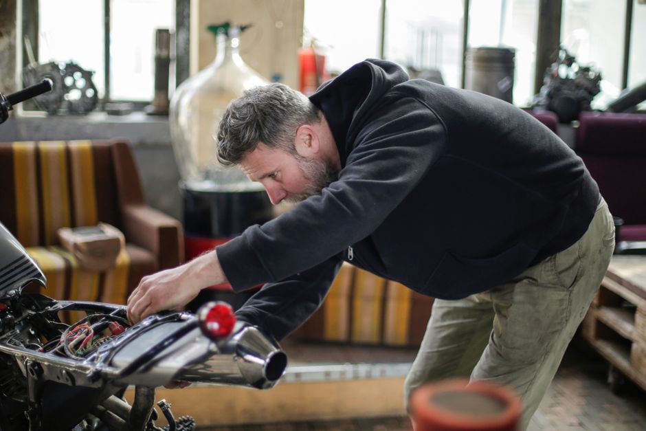 Side view of serious bearded adult male mechanic in black workwear checking electric details of motorbike while working in spacious garage